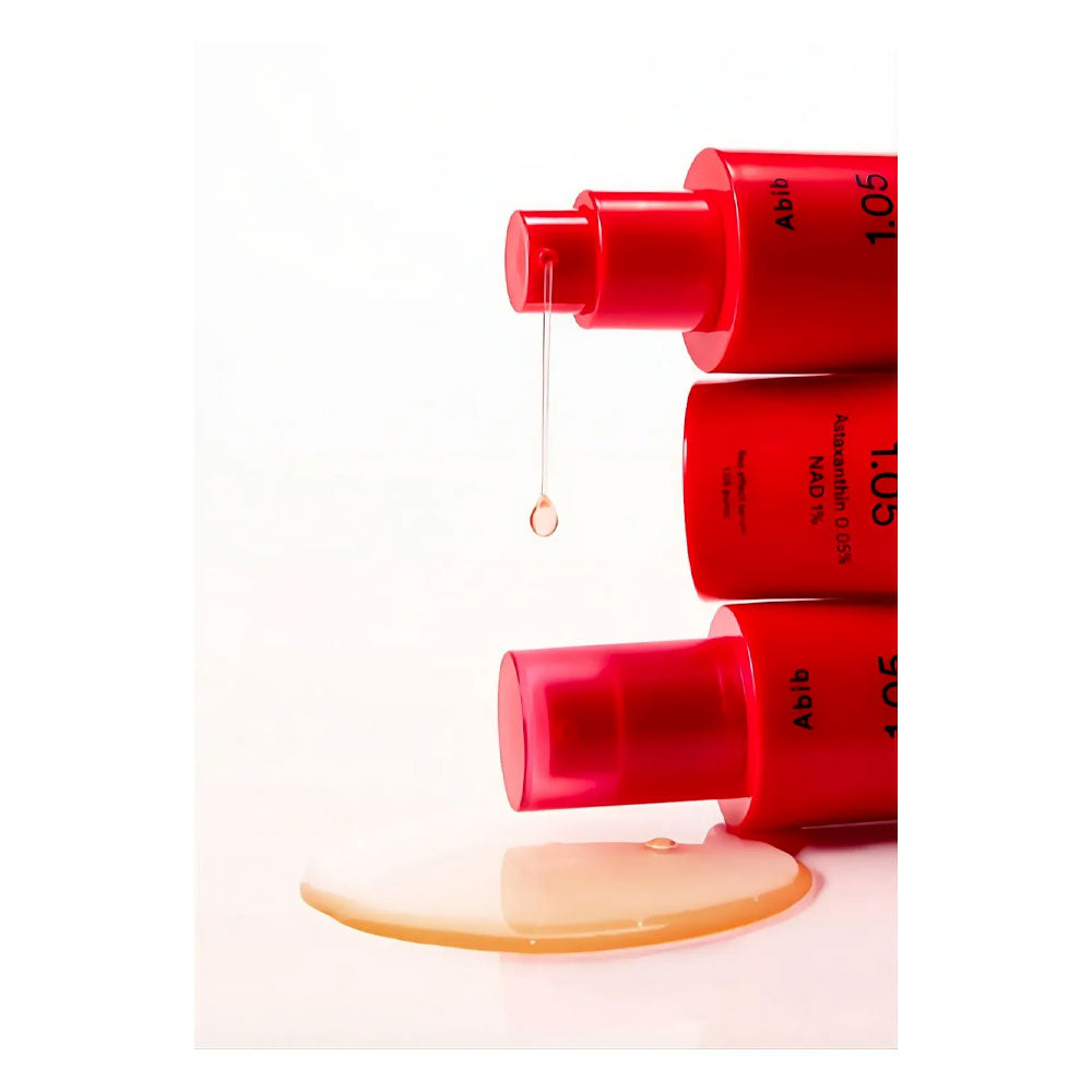 Three red cosmetic pump bottles stacked vertically on a white background, with a single drop of clear liquid hanging above and a pool of pale golden serum below.