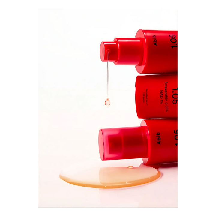 Three red cosmetic pump bottles stacked vertically on a white background, with a single drop of clear liquid hanging above and a pool of pale golden serum below.