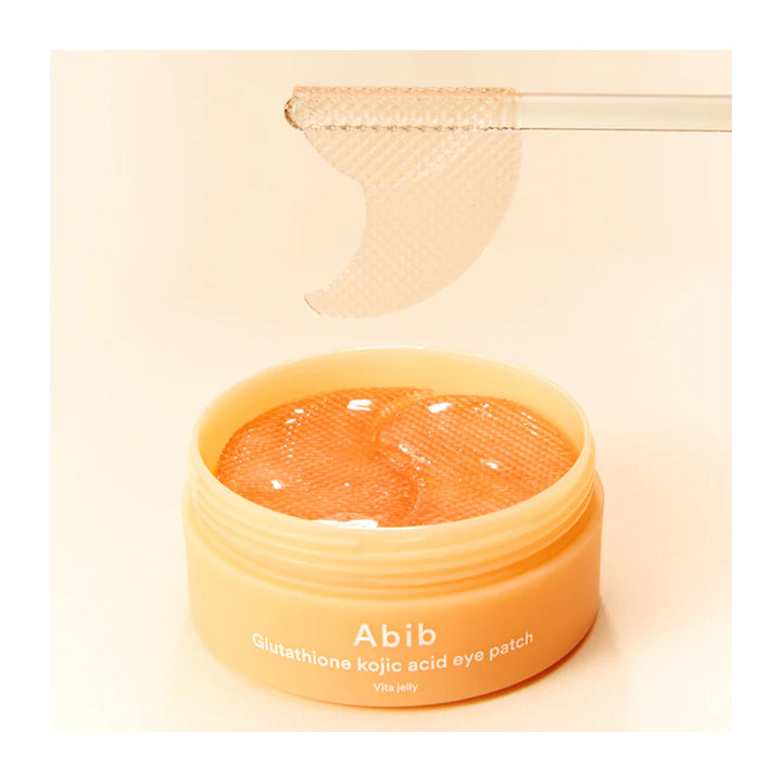 An open peach-colored jar of Abib Glutathione Kojic Eye Patch showing the orange gel inside, with a single crescent-shaped eye patch floating above on a clear stick against a warm cream background.