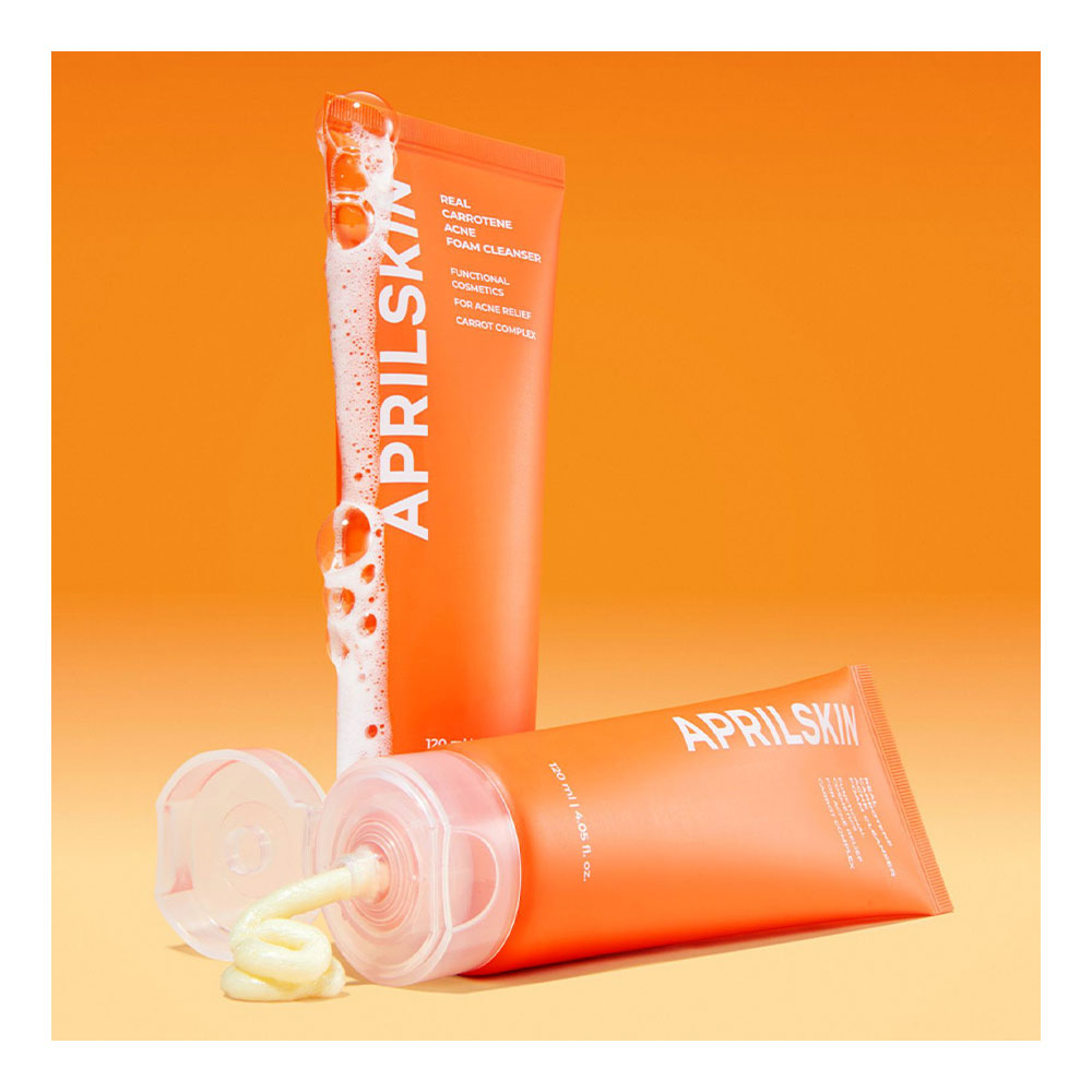 Aprilskin face cleansing foam in an orange squeeze tube, shown standing upright with white foam dripping from the top and another tube lying on its side with the cap off and some foam visible. The background is a clean orange gradient.