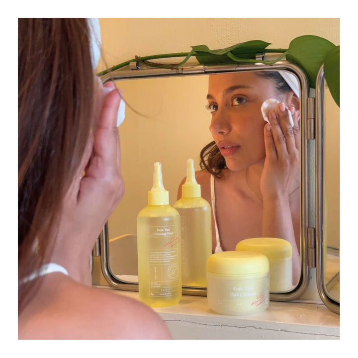 A woman applies skincare cream to her face while looking into a mirror. In the foreground, three yellow skincare bottles and two cream jars are arranged on a bathroom counter.