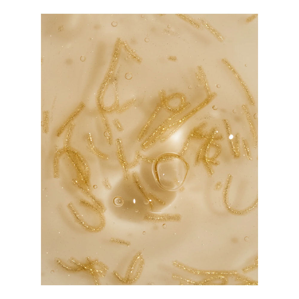 A close-up of a beige or cream-colored liquid surface with scattered fine gold glitter strands and a few small clear bubbles.