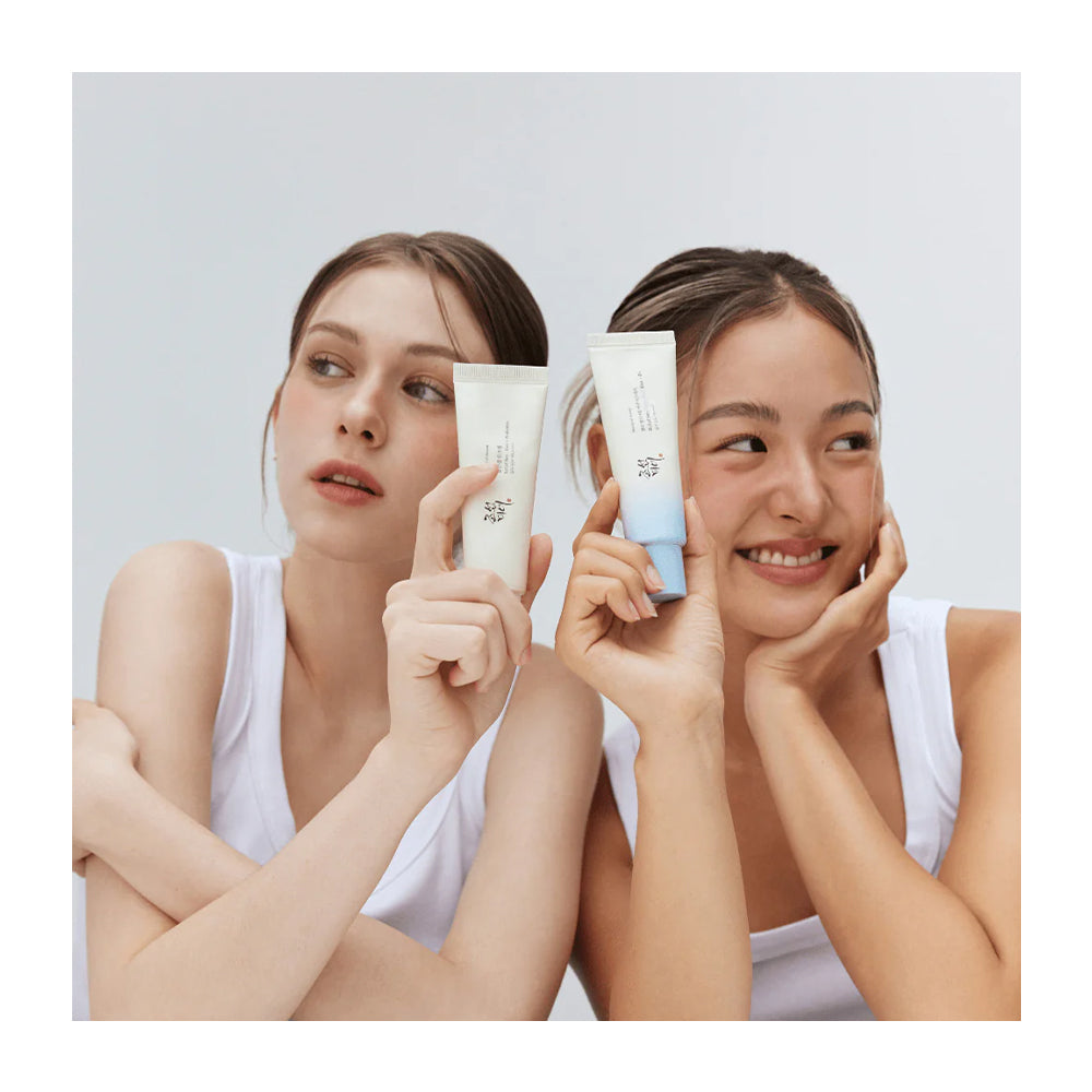 Two women in white tank tops hold up skincare tubes toward the camera against a light gray studio background.