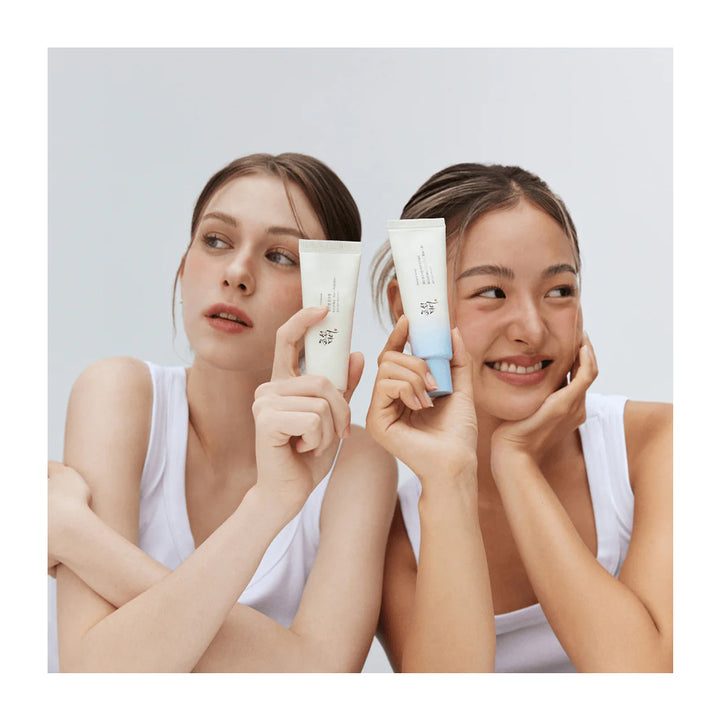 Two women in white tank tops hold up skincare tubes toward the camera against a light gray studio background.
