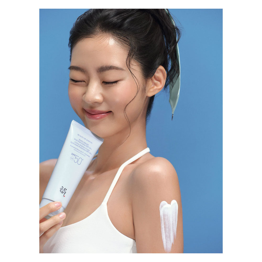 A woman with dark hair tied back wears a white camisole and has white sunscreen lotion applied to her upper arm. She is holding a white tube of sunscreen against a solid blue background.