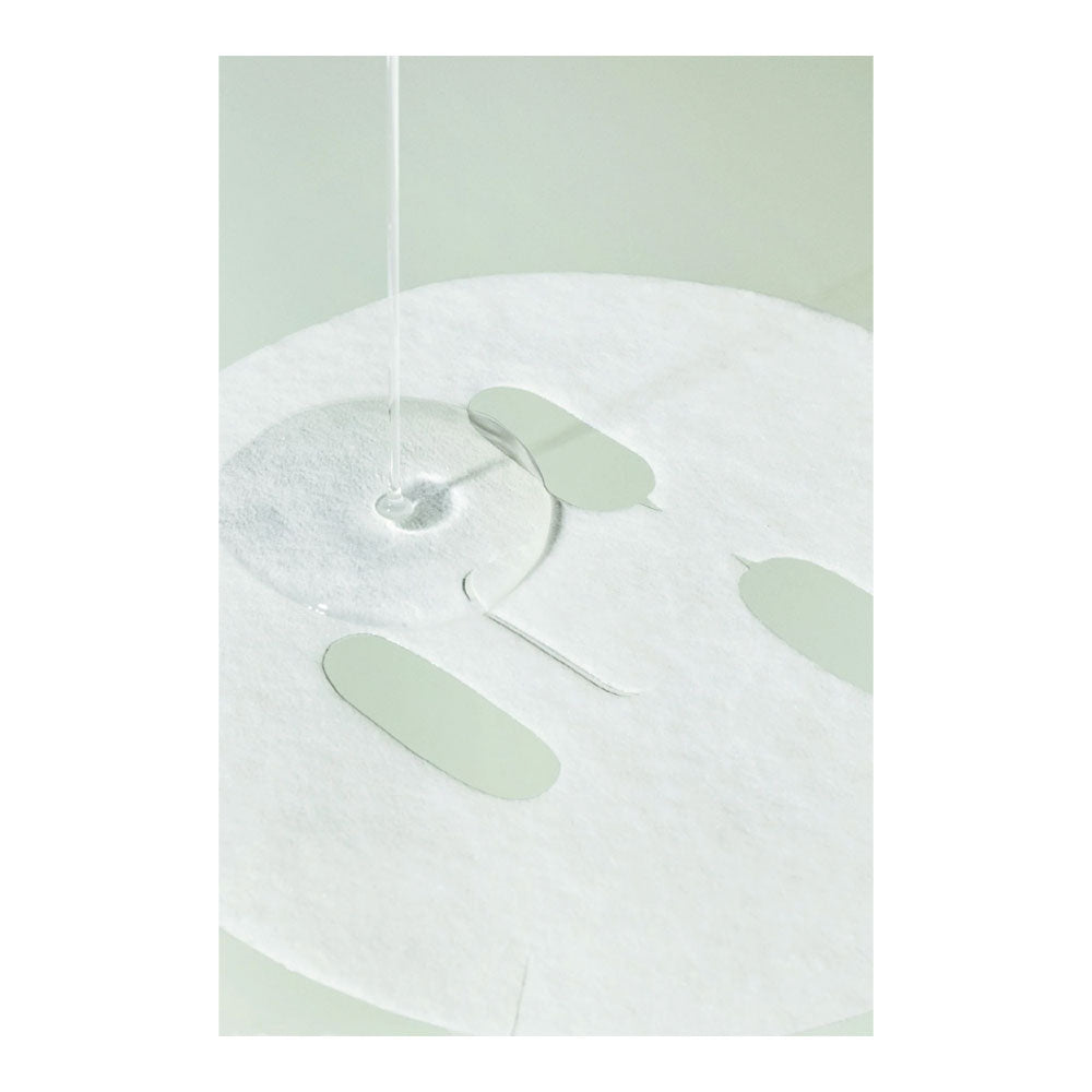 A close-up of a white sheet mask with cutout openings, showing a clear liquid being poured into one of the circular openings, with pale mint-green liquid droplets visible on the surface.