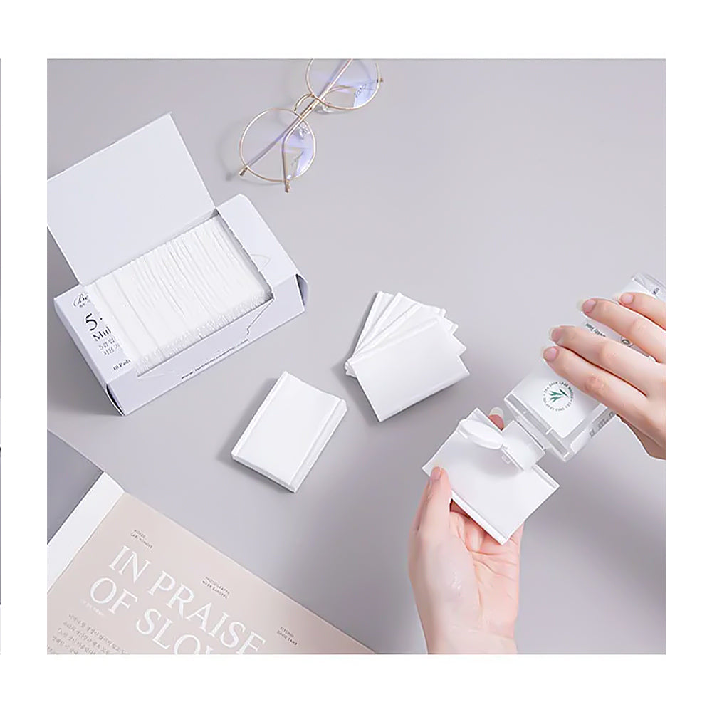 A top-down view of a person's hands opening a white box of square cotton pads and dispensing them onto a light gray surface, with a pair of round glasses and a book also visible on the tabletop.