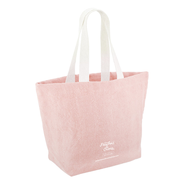 A light pink canvas tote bag with white shoulder straps and white printed text near the bottom right, shown on a plain white background.