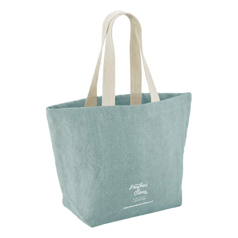 A light blue-green canvas tote bag with natural beige fabric shoulder straps and white script branding on the lower front, shown against a plain white background.