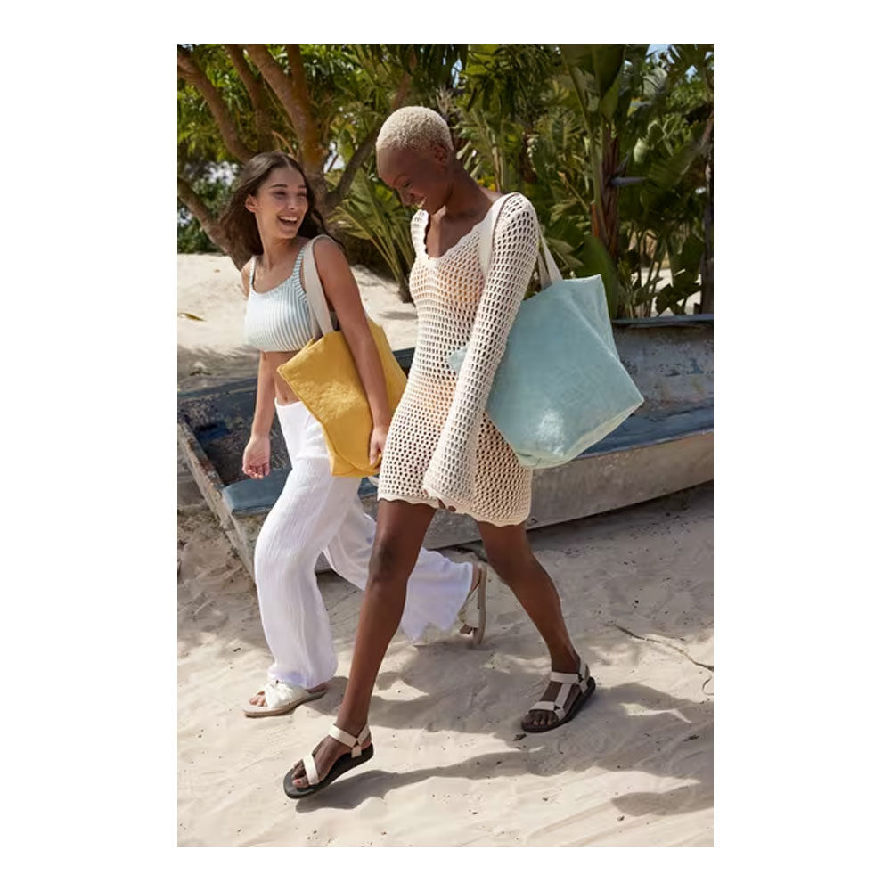 Two women walking on a sandy beach wearing crochet knit clothing and carrying tote bags. One woman wears a cream crochet mini dress and black sandals, holding a light blue tote bag, while the other wears a white ribbed tank top and white pants, holding a yellow bag.