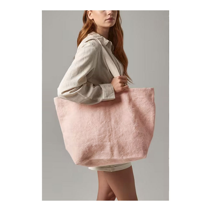 A woman wearing a light beige linen shirt and shorts carries a large, soft blush pink fuzzy fabric tote bag against a solid gray studio background.