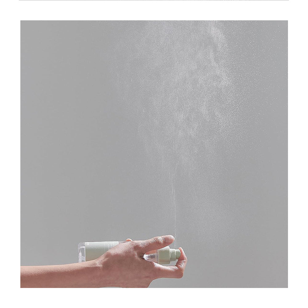 A hand holding a clear spray bottle with a white pump, spraying a fine mist upward against a plain light gray background.