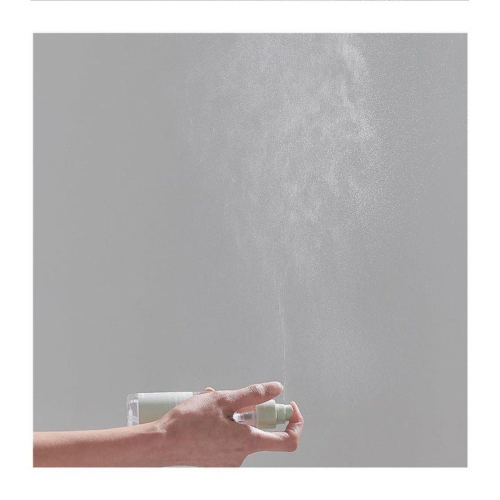 A hand holding a clear spray bottle with a white pump, spraying a fine mist upward against a plain light gray background.