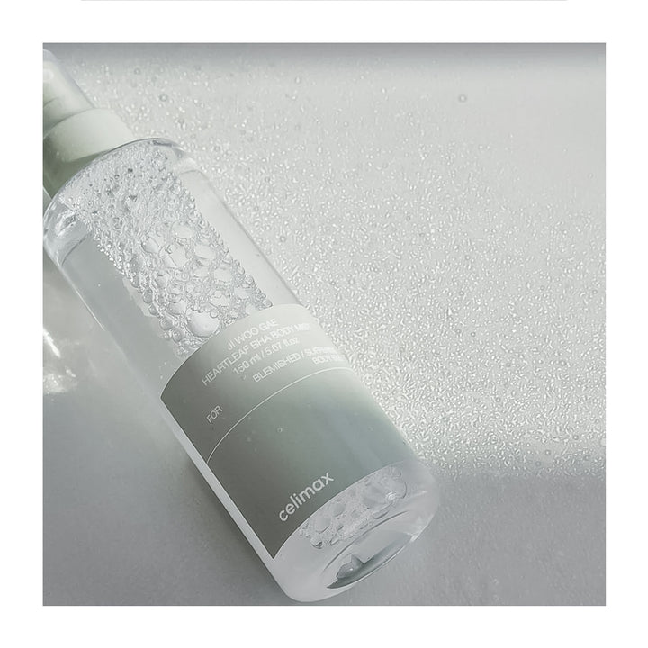 A clear cylindrical bottle of Celimax cleansing liquid with a white pump, lying diagonally on a textured light gray surface with water droplets.