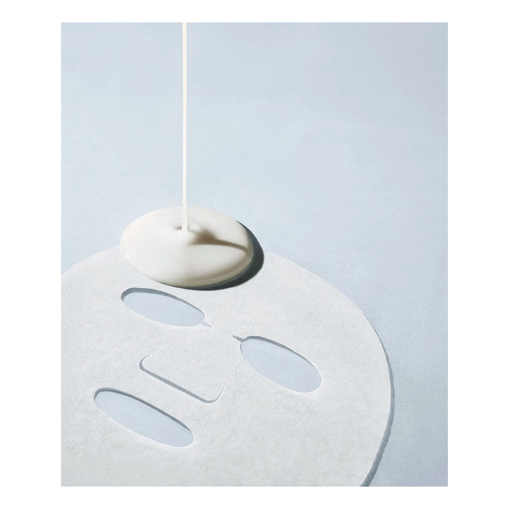 A white sheet mask with cutout openings lies on a pale blue background, with a single vertical droplet of white cream or serum dripping onto the center of the mask.