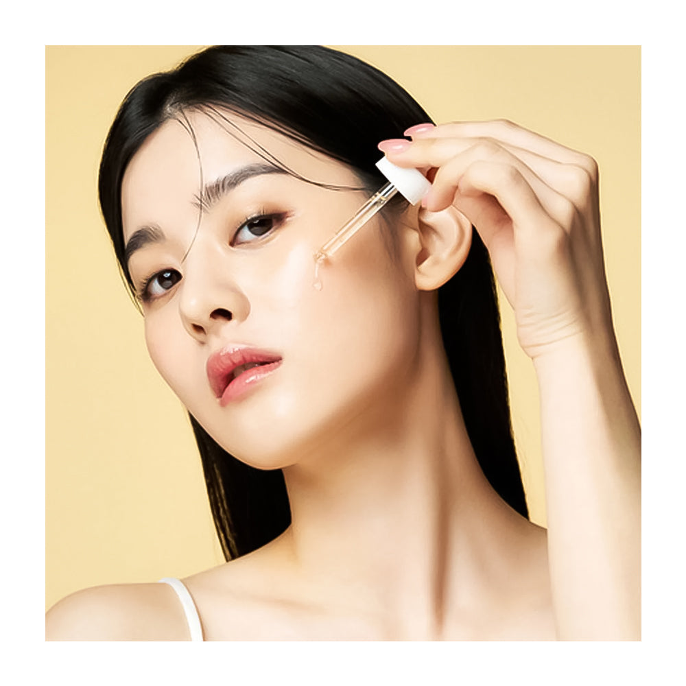 A close-up of a woman with long black hair applying a skincare serum or dropper cream to her cheek against a warm beige background.