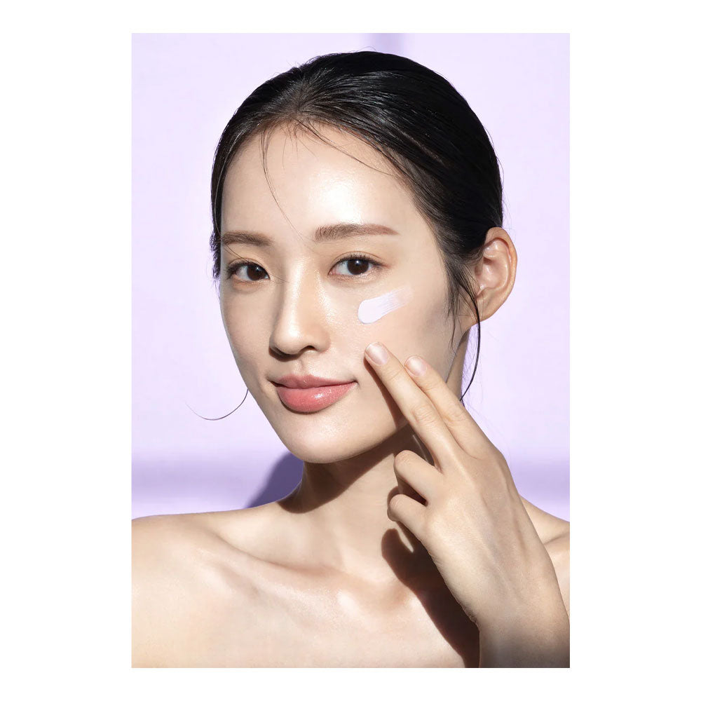 A close-up of a woman with dark hair pulled back, applying or spreading a white cream product on her cheek against a soft light purple background.