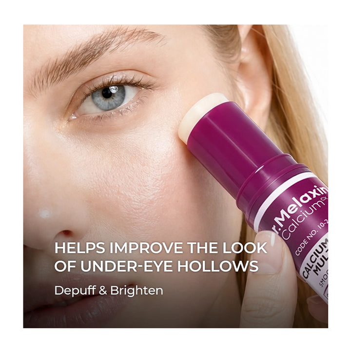 Close-up of a woman applying an under-eye cream stick to her cheek. The product is a purple and white tube labeled &
