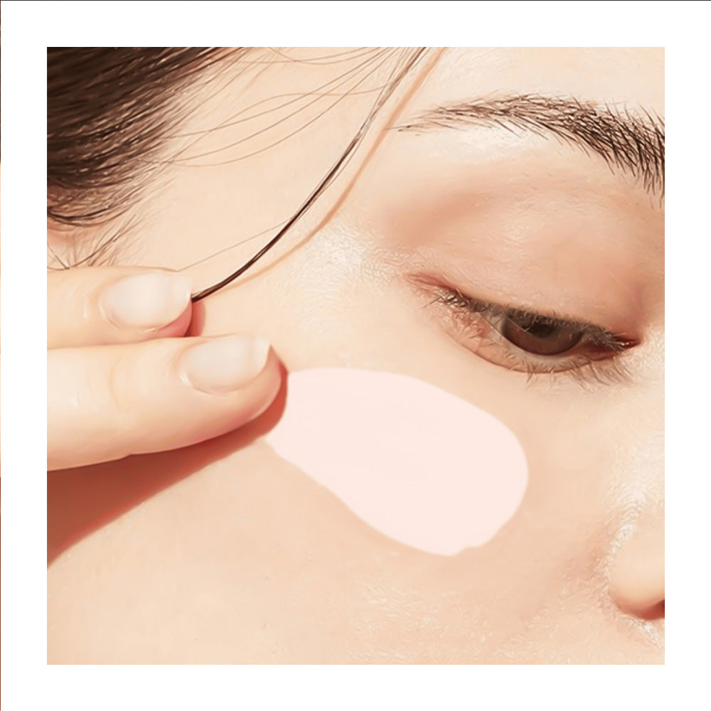 Close-up of a person's face with a pale pink makeup swatch or patch applied to the cheek, held in place by a finger.