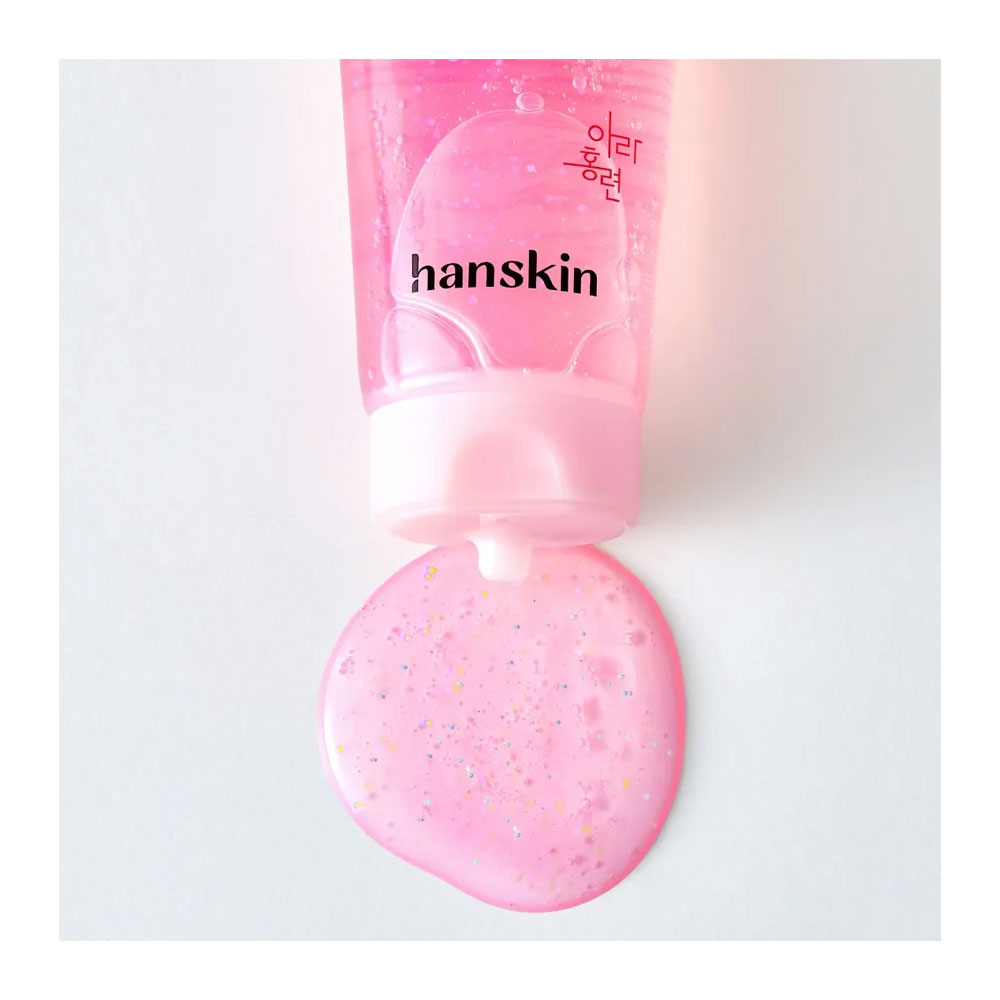 A pink Hanskin cosmetic tube lying on its side with a dollop of translucent pink gel and multicolor glitter being squeezed out onto a light gray background.