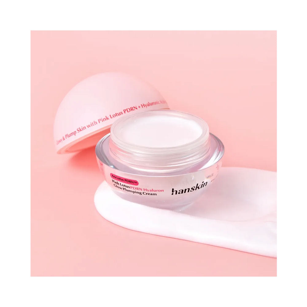 A jar of hanskin Pump Skin with Pink Lotus PDRN + Hyaluronic Acid Pumping Cream, shown open with its pink lid leaning behind it and a thick smear of the white cream in front, set against a solid pink background.