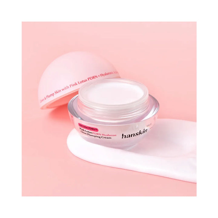 A jar of hanskin Pump Skin with Pink Lotus PDRN + Hyaluronic Acid Pumping Cream, shown open with its pink lid leaning behind it and a thick smear of the white cream in front, set against a solid pink background.