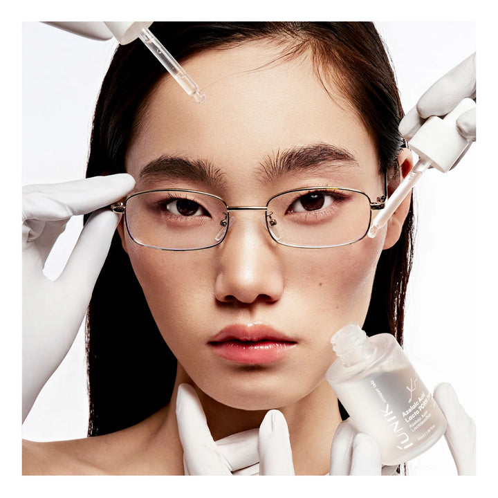 A close-up beauty shot of a model with clear liquid being applied to her face using glass dropper bottles and white-gloved hands. She is wearing thin metal-framed rectangular eyeglasses against a clean white background.