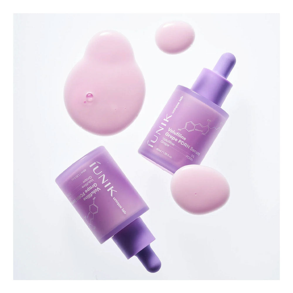 Two bottles of iUNIK Grape Enzyme Serum in a frosted lavender purple color with matching droppers, arranged on a white background with decorative pink liquid droplets.