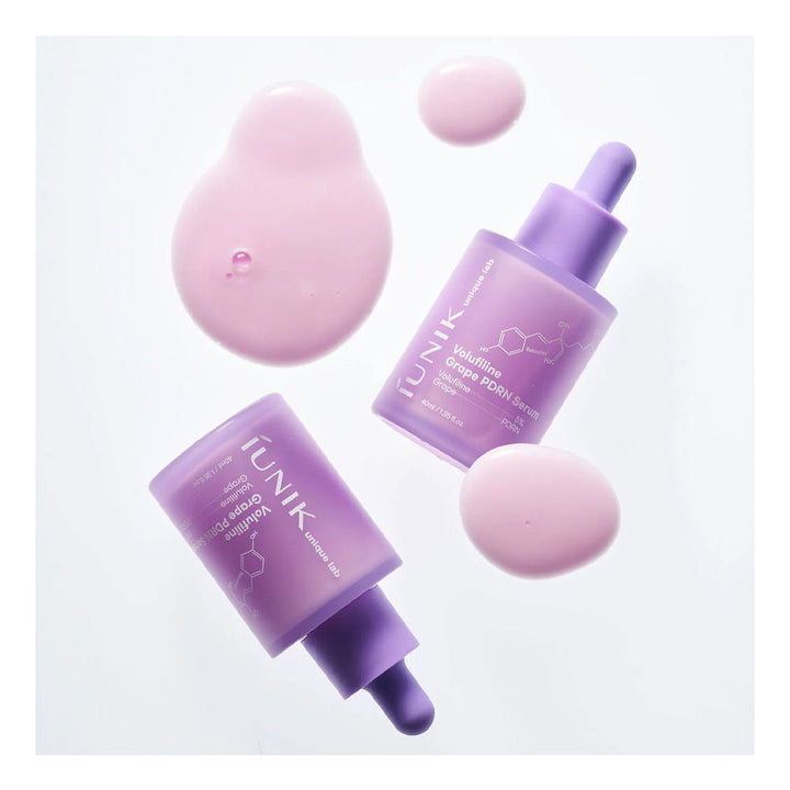 Two bottles of iUNIK Grape Enzyme Serum in a frosted lavender purple color with matching droppers, arranged on a white background with decorative pink liquid droplets.