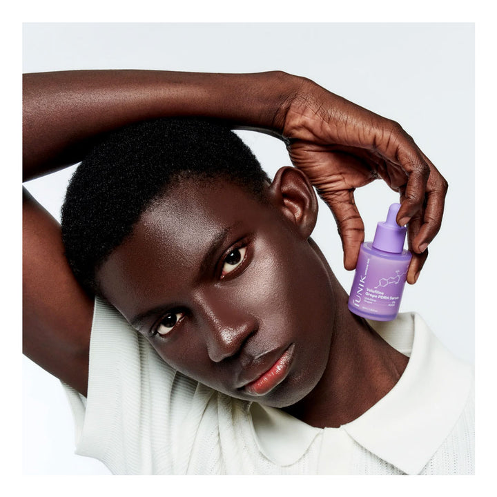 A close-up lifestyle shot of a model with dark skin and short black hair, wearing a white ribbed polo shirt and holding a small purple bottle of MAC Cosmetics Prime Time Radiance Serum against a light gray background.