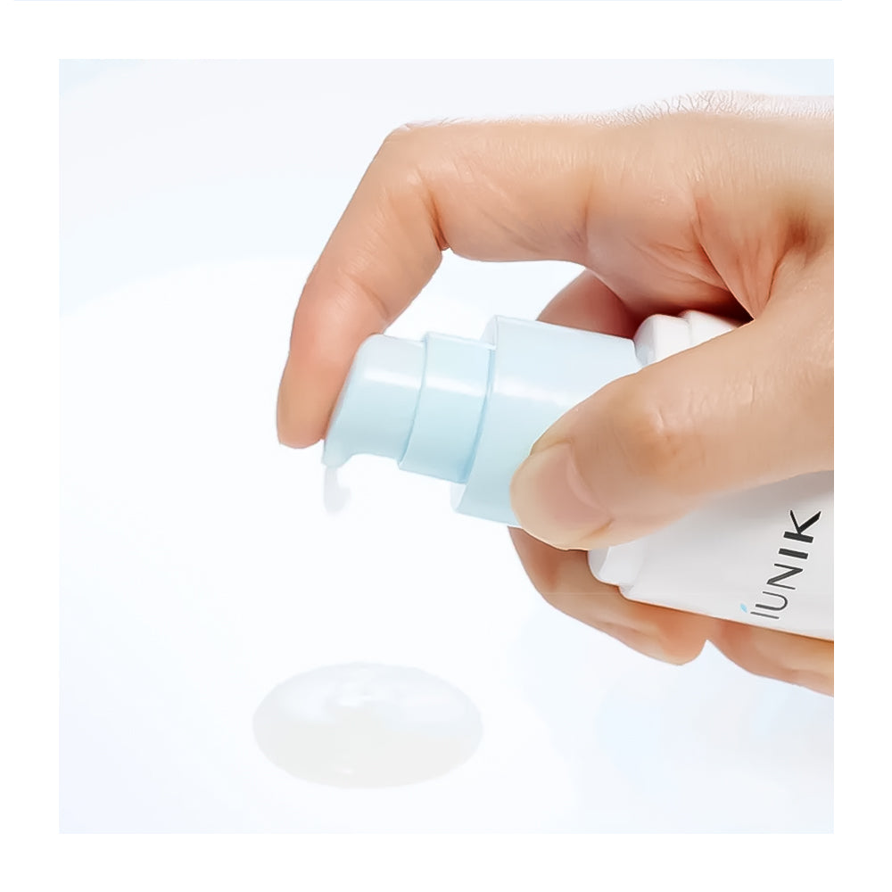 A close-up of a hand holding a white skincare pump bottle with a pale blue pump, dispensing a clear gel or cream onto a white surface. The brand name &