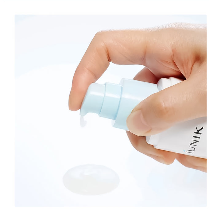 A close-up of a hand holding a white skincare pump bottle with a pale blue pump, dispensing a clear gel or cream onto a white surface. The brand name &