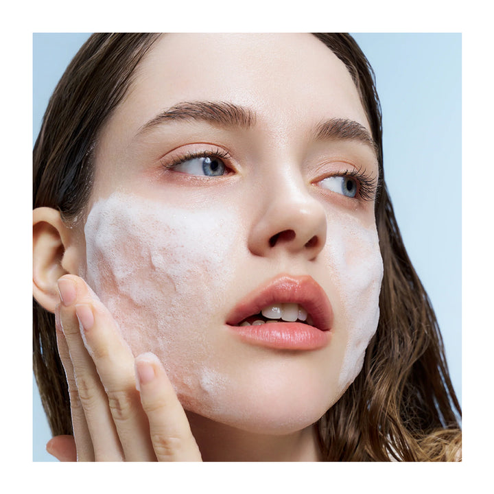 Close-up of a woman with white foaming cleanser on her face, touching her cheek, against a light blue background.