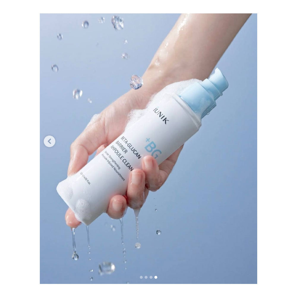 A hand holding a white and light blue bottle of IUNIK +BG Hair Glucon + Mousse Clean against a light blue background with water droplets.
