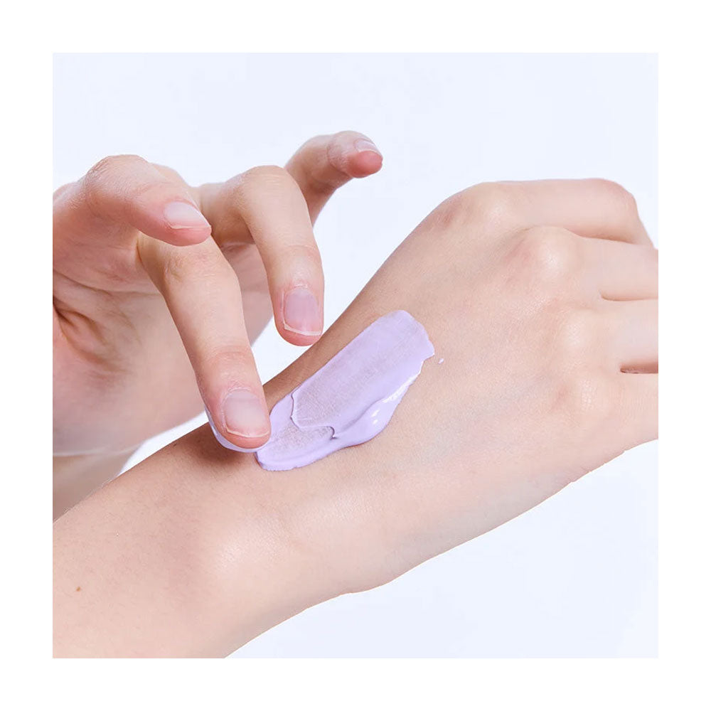 A close-up of a hand applying or spreading a pale lavender cosmetic cream or gel onto the back of another hand against a clean white background.