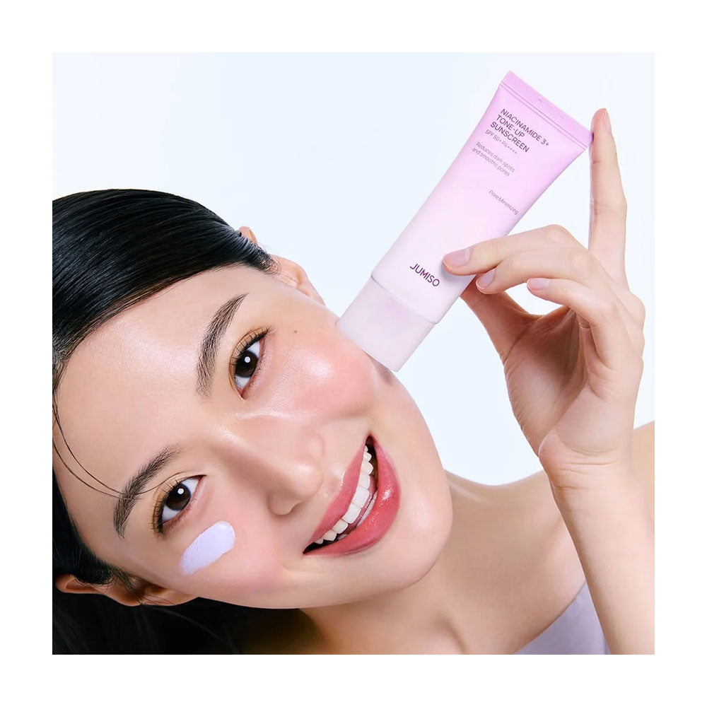A woman with a radiant complexion holds a pink tube of Cover Up Moisturizing Face Cream next to a dollop of the cream on her cheek against a clean white background.