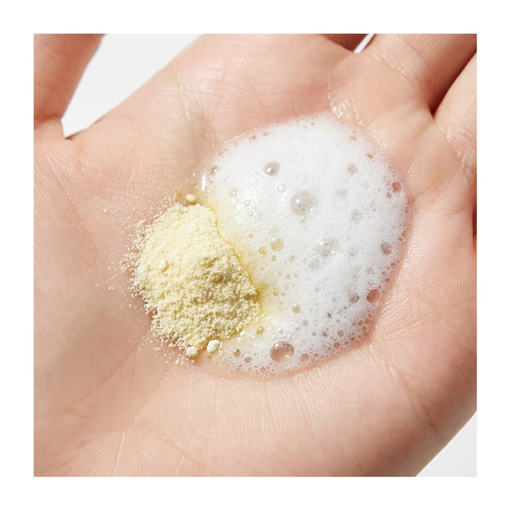 A close-up of a human hand holding a small pile of pale yellow powder next to a pool of white foam.
