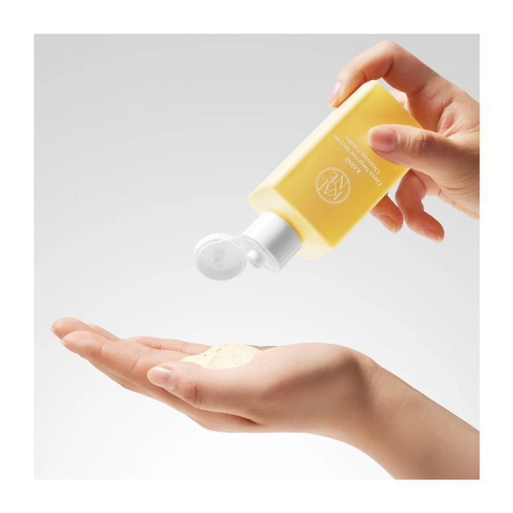 A hand holds a yellow bottle and dispenses a small amount of creamy beige product onto the palm of another hand, shown against a light gray studio background.