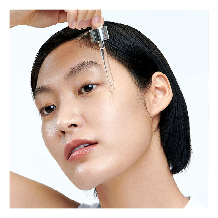A close-up of a woman with wet, dewy skin applying a clear serum dropper from a glass pipette to her forehead, with a hand holding the applicator above her head against a clean white background.