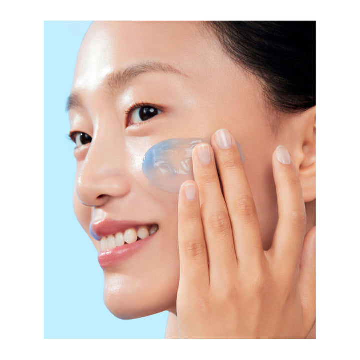 A close-up of a woman with a translucent blue gel or patch applied to her cheek, touching her face with her hand against a light blue background.