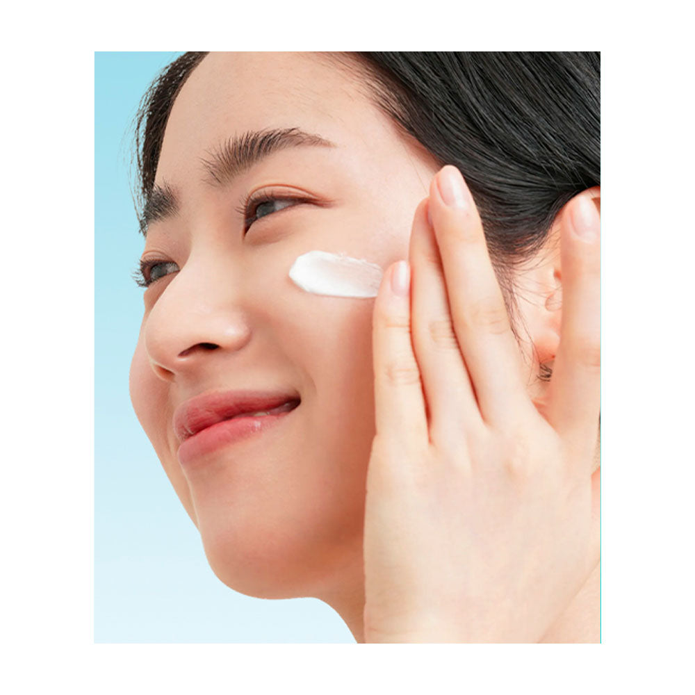Close-up of a woman applying white cream to her cheek against a light blue background.