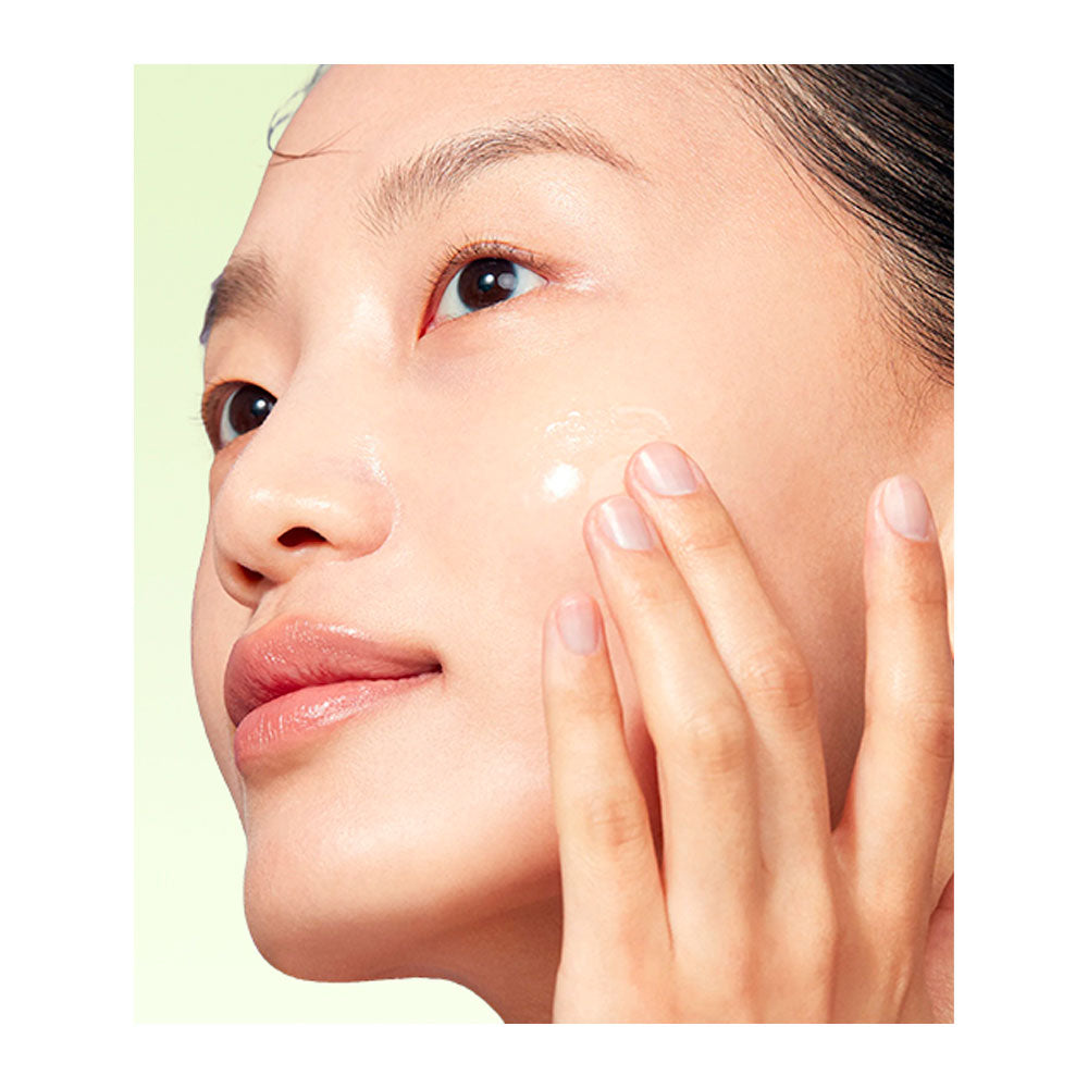 Close-up of a woman applying a clear skincare cream to her cheek, with her hand gently touching her face against a pale green background.