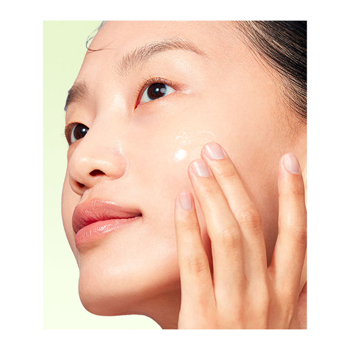 Close-up of a woman applying a clear skincare cream to her cheek, with her hand gently touching her face against a pale green background.