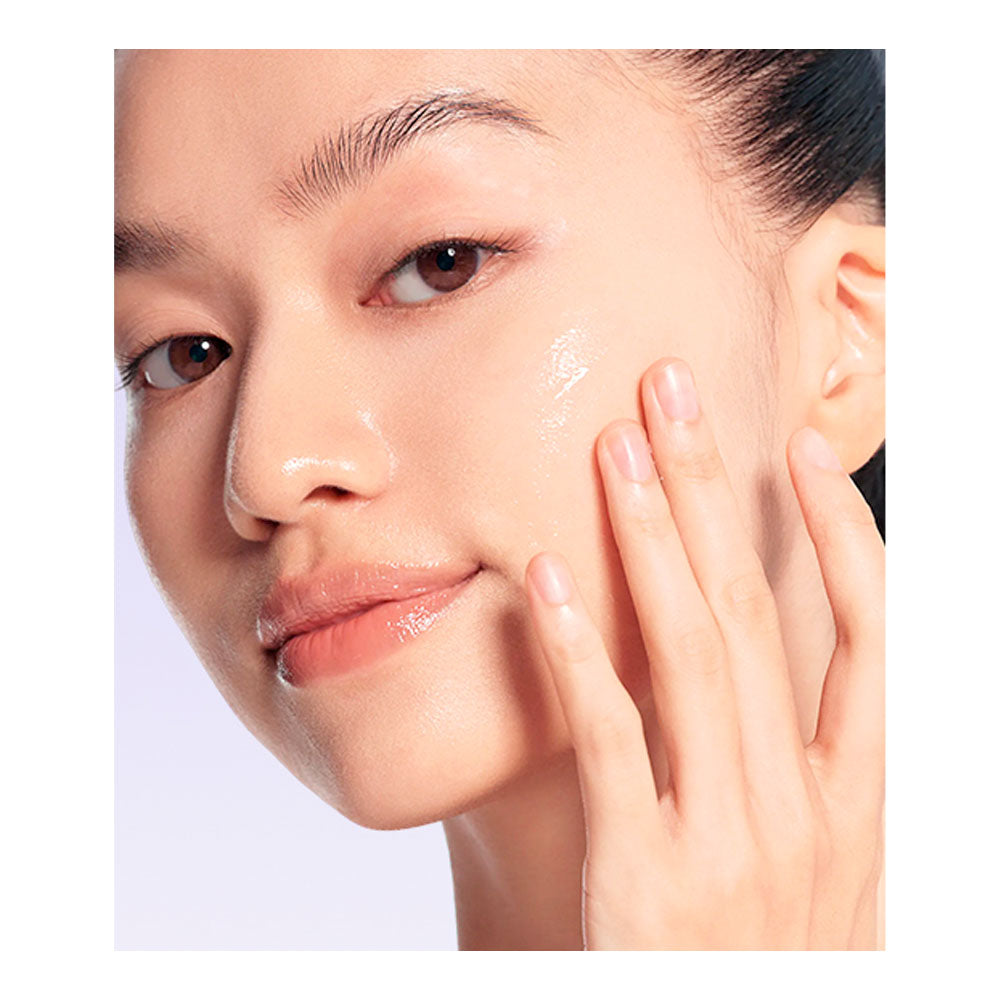Close-up of a woman's face and hand applying a glossy, luminous skincare cream or serum to her cheek against a clean, light background.