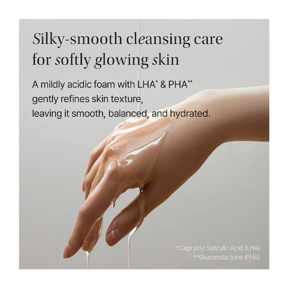 A hand and forearm covered with a glossy, silky cleansing cream, set against a soft light gray background with descriptive text about soothing, mildly acidic foaming care.