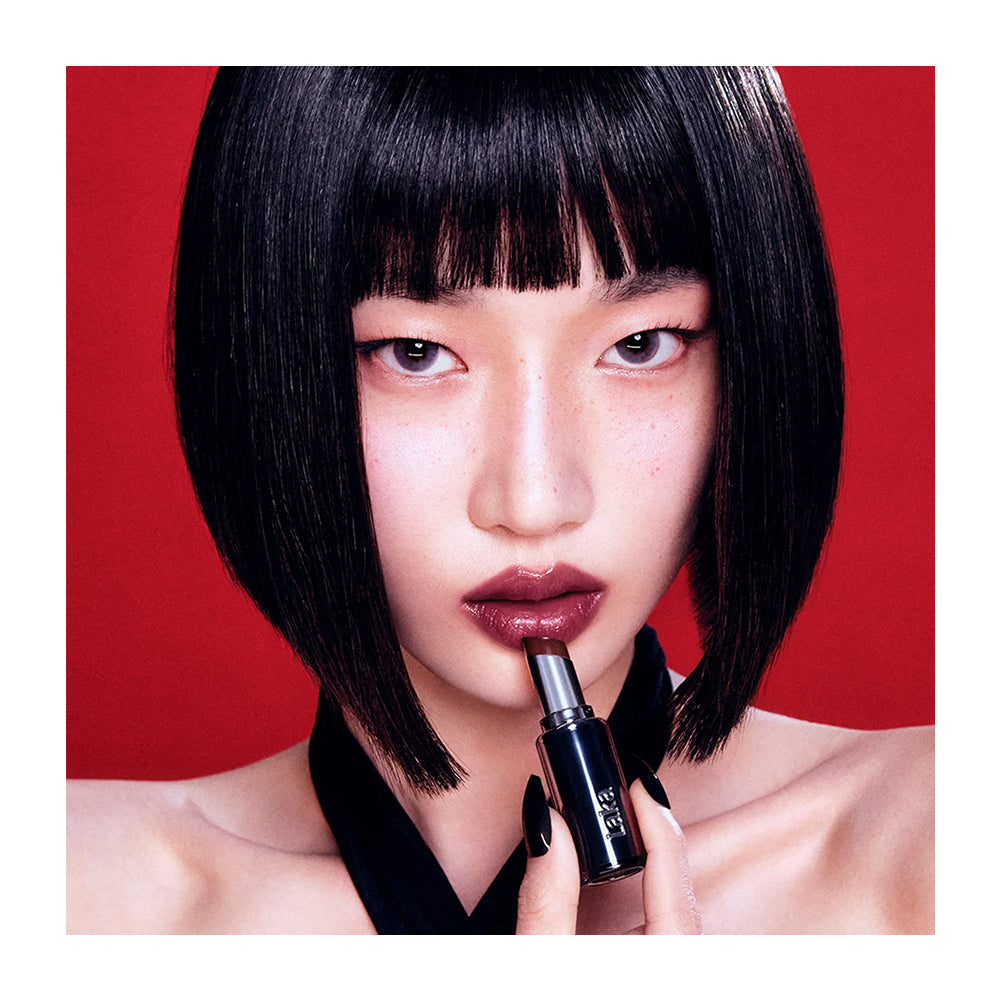 A close-up portrait of a model with a black bob haircut applying a dark brown lipstick against a solid red background.