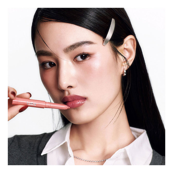 A close-up of a woman with long black hair, wearing a white collared shirt and gray cardigan, holding a pink lip liner pencil near her lips against a clean white background.
