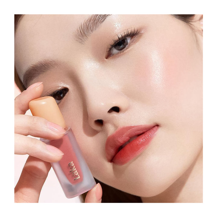 A close-up of a woman holding a frosted lip product tube near her face, showing a glossy peach-pink lip color. The brand name &