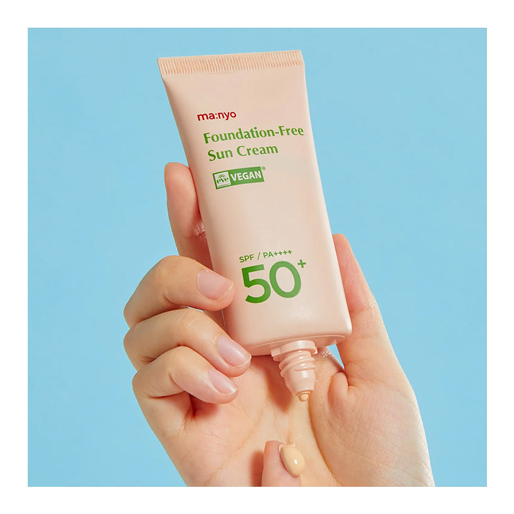 A hand holding a squeeze tube of ma-nyo Foundation-Free Sun Cream SPF 50+ against a solid light blue background. The tube is a pale peach-beige color with green and red text, and a small amount of the beige sunscreen cream is visible dripping from the tip.