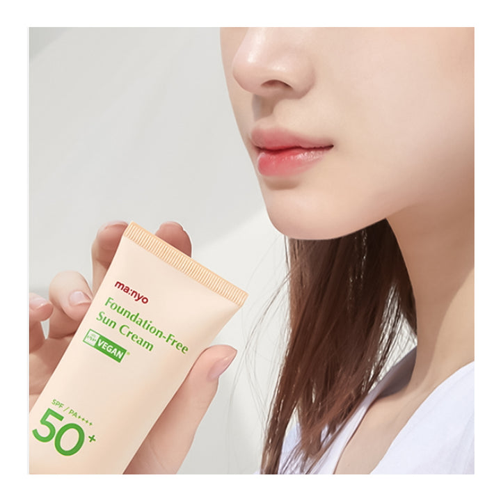 A close-up lifestyle shot of a woman holding a squeeze tube of ma:nyo Foundation-Free Sun Cream SPF 50+ with PA++++. The tube is a soft peach-beige color with green and red text, and the woman&
